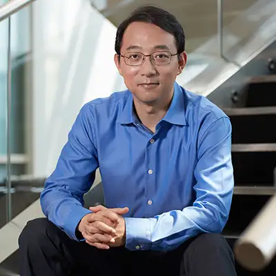 Man in a blue shirt and glasses seated in a modern office setting, with a staircase in the background.