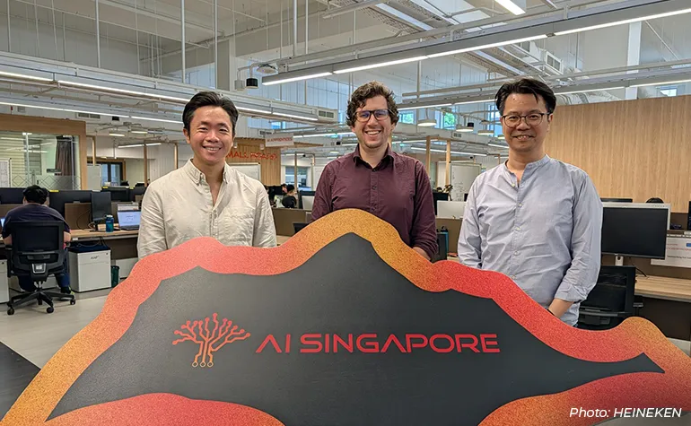 Three team members stand smiling behind an “AI Singapore” sign inside a modern open-plan office, with workstations and employees visible in the background.
