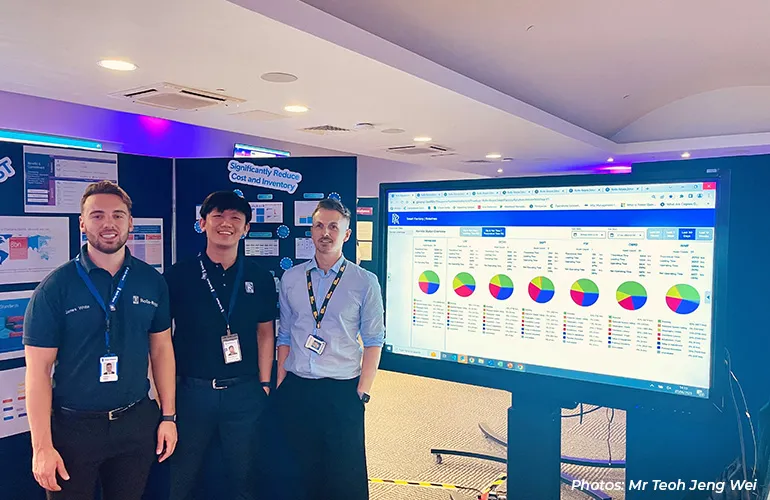 Three men stand beside a large screen displaying colorful data charts at a technology showcase.