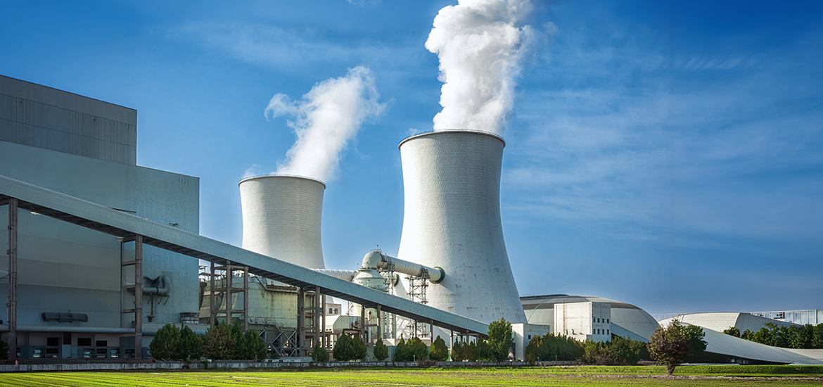 Industrial facility with large cooling towers emitting steam, representing energy generation or heavy industrial operations, set against a clear blue sky and surrounded by supporting infrastructure and green landscaping.