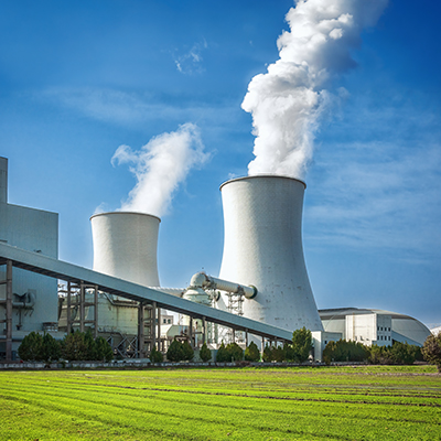 Industrial facility with large cooling towers emitting steam, representing energy generation or heavy industrial operations, set against a clear blue sky and surrounded by supporting infrastructure and green landscaping.