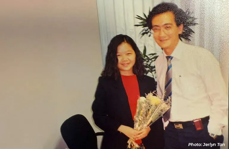 Two people posing indoors, with the woman holding a bouquet of flowers while standing beside a man in a shirt and tie.