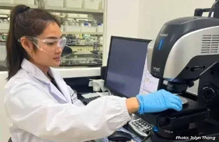 Scientist wearing safety goggles and gloves operating laboratory equipment beside a computer in a lab setting.