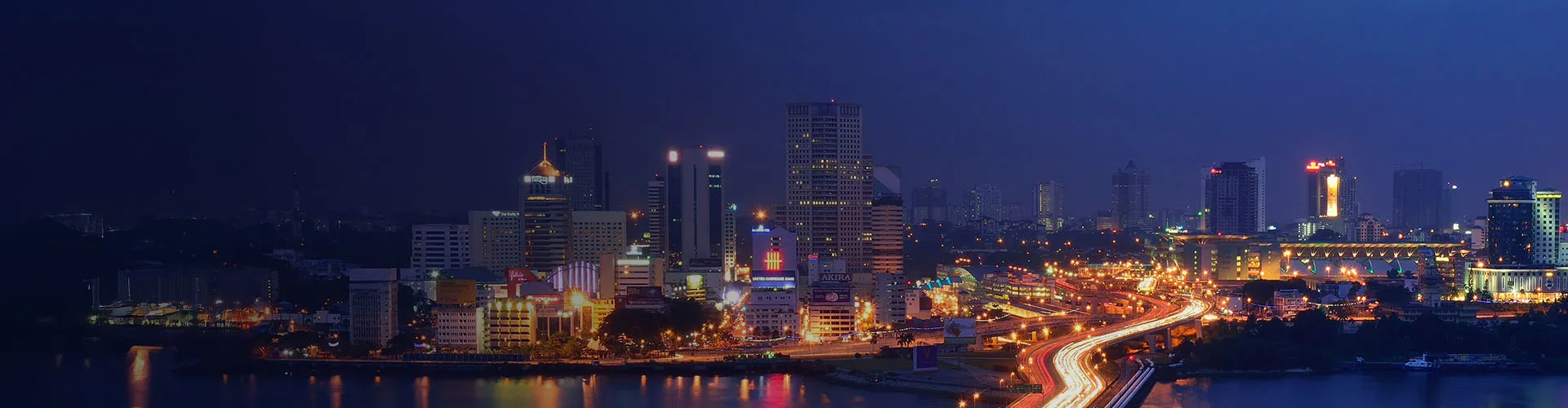 City skyline at night with illuminated skyscrapers and light trails from vehicles on a busy highway.