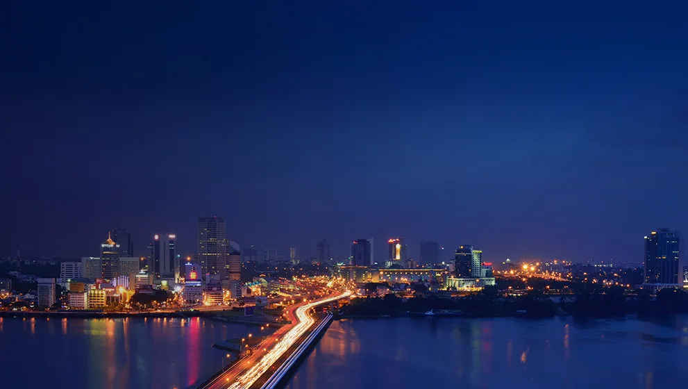 City skyline at night with illuminated skyscrapers and light trails from vehicles on a busy highway.