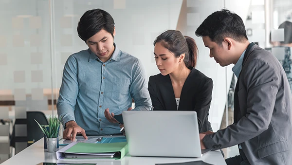 Three professionals having a discussion around a laptop and documents in a modern office setting.
