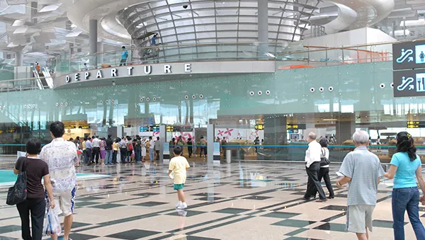 Travelers walking through the spacious departure hall of a modern airport, with glass architecture, high ceilings, and check-in counters in the background.
