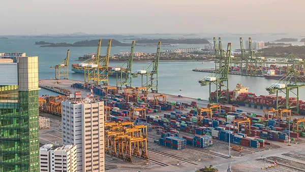 Aerial view of a busy shipping port in Singapore, featuring numerous colorful cargo containers, large container cranes, and nearby modern buildings, set against a coastal landscape.