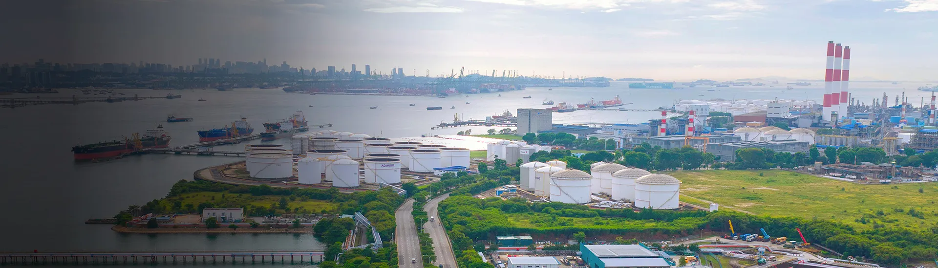 Aerial view of a coastal industrial area with oil storage tanks, port facilities, ships, and power plant chimneys along the waterfront.