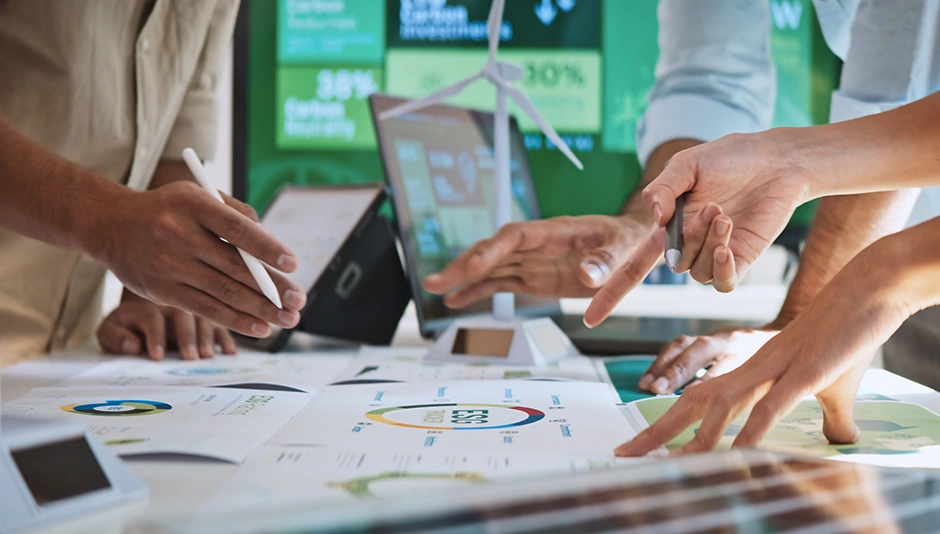 Team collaborating on renewable energy plans with charts, digital tablets, and a miniature wind turbine on the table.