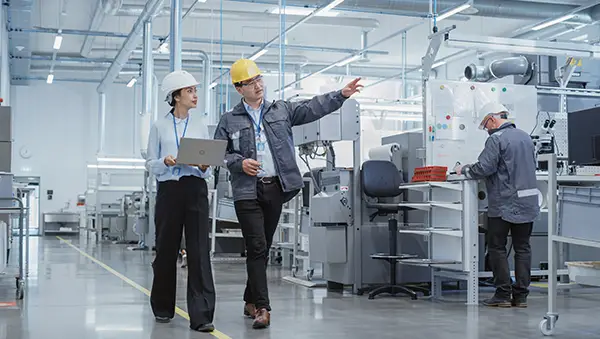 Two engineers in hard hats and ID lanyards walk through a modern, brightly lit manufacturing facility, discussing operations while another technician works in the background—symbolizing innovation and industrial collaboration.