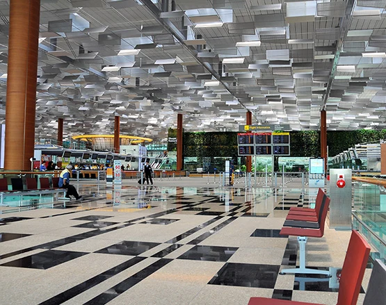 Spacious and modern airport terminal with geometric ceiling design, red seating, digital flight information boards, and a few travelers walking through the departure area.
