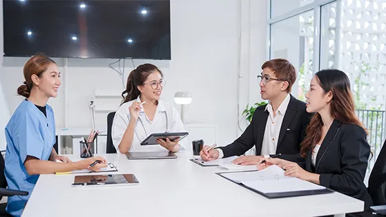 Healthcare professionals and business leaders seated around a meeting table in a bright office, discussing ideas with tablets and documents during a collaborative strategy meeting.