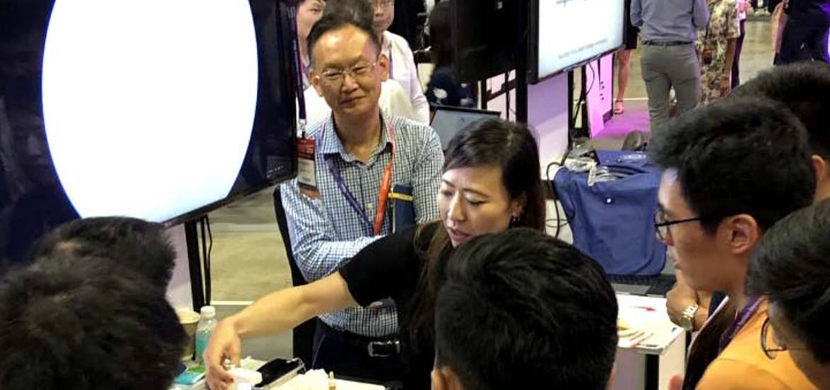  group of people gathers around a demonstration booth at a tech or trade exhibition. A woman presenter is actively explaining or showcasing a small medical or technical device, while the attendees listen attentively. Monitors, lanyards, and booths are visible in the background.
