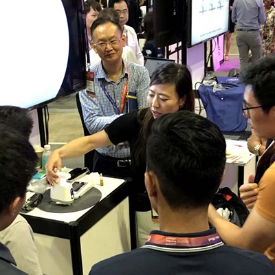  group of people gathers around a demonstration booth at a tech or trade exhibition. A woman presenter is actively explaining or showcasing a small medical or technical device, while the attendees listen attentively. Monitors, lanyards, and booths are visible in the background.