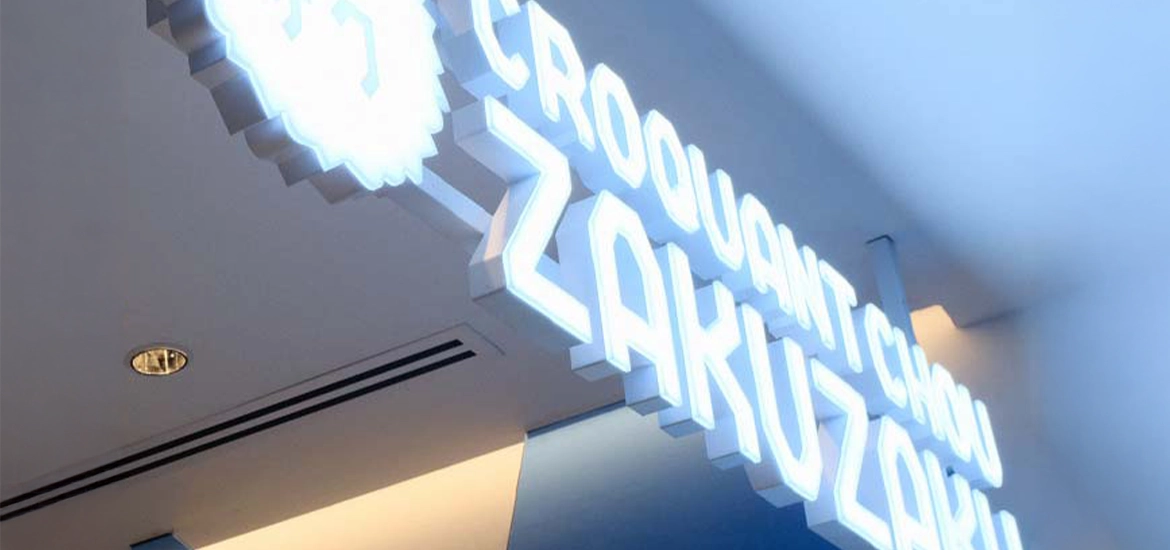 Illuminated store sign of "Croquant Chou Zakuzaku" mounted on a ceiling. The bold, glowing letters are styled in a modern, geometric font, creating a clean and eye-catching effect. The overall ambiance is cool-toned with soft blue lighting.