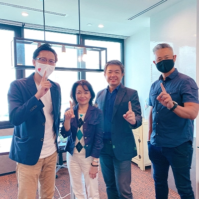 Four people standing indoors, smiling and holding up one finger each in a gesture of unity or achievement; two are wearing face masks, and all are dressed in business casual attire.