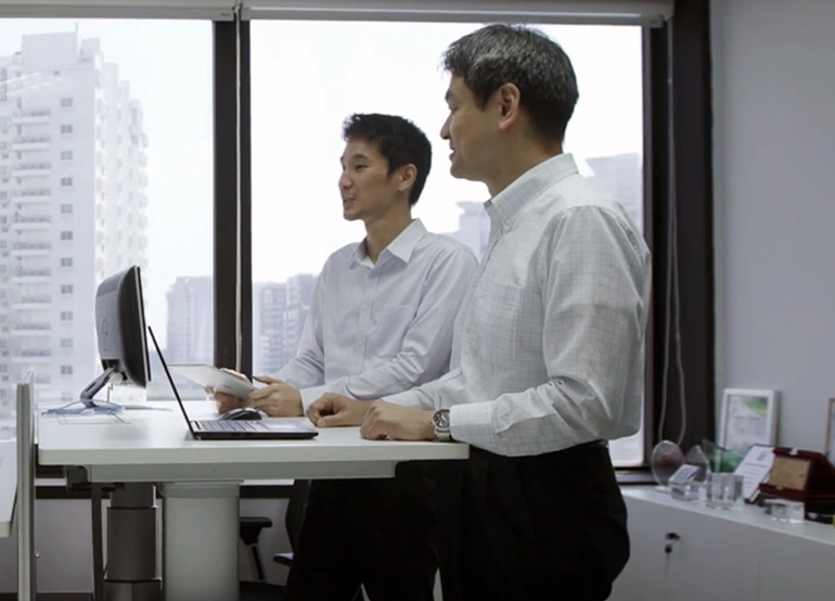 Two Asian businessmen in formal white shirts stand at a height-adjustable desk in a modern office with large windows overlooking a city skyline. One works on a laptop while the other looks at a desktop monitor, suggesting a collaborative and professional work environment.