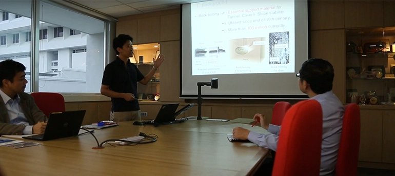 A business meeting or technical presentation in a modern office setting, featuring a presenter standing and pointing at a projection screen displaying slides on rock bolting. Three seated professionals attentively watch, taking notes or working on laptops, in a well-lit room with large windows and a backdrop of display cabinets.