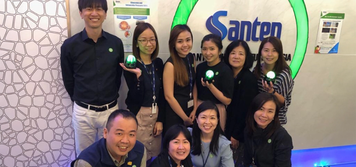 Group photo of smiling employees at Santen Pharmaceutical Asia in front of a company sign. They are holding green-lit objects and standing in a well-lit, modern reception area, suggesting a celebratory or awareness campaign.