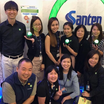 Group photo of smiling employees at Santen Pharmaceutical Asia in front of a company sign. They are holding green-lit objects and standing in a well-lit, modern reception area, suggesting a celebratory or awareness campaign.
