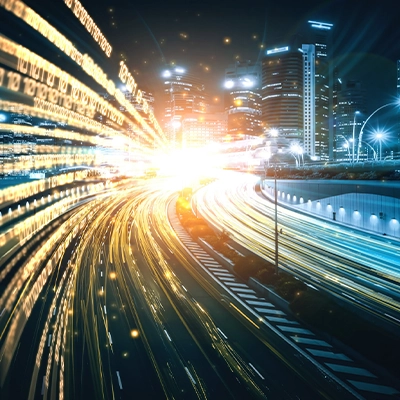 A futuristic cityscape at night with illuminated skyscrapers and highways filled with glowing light trails. Binary code streams through the air, symbolizing data and digital connectivity, converging toward a bright light source in the center.