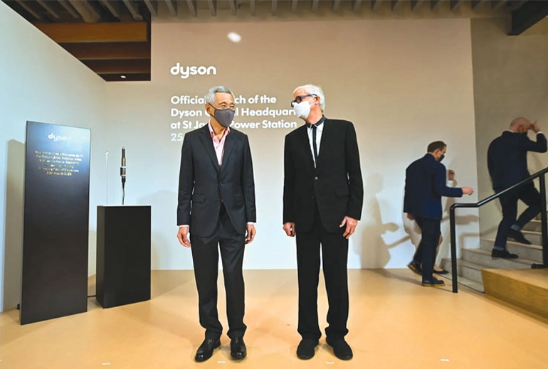Two men in formal attire wearing face masks stand in front of a Dyson display at the official launch of the Dyson Global Headquarters at St James Power Station on March 25. The event setting features clean, minimalist design with additional guests in the background.