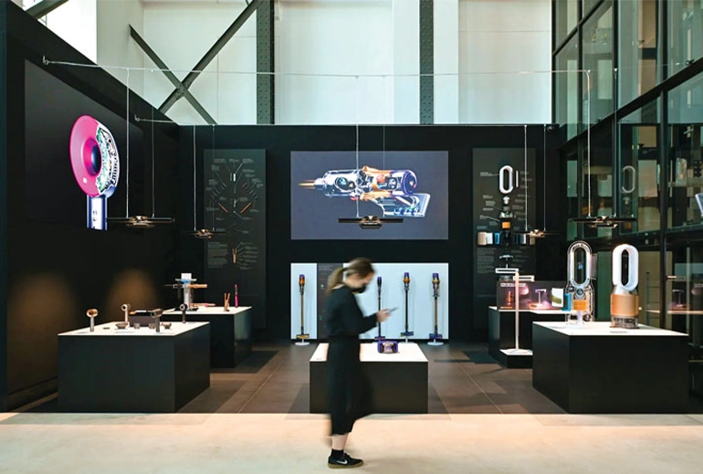 A woman walks past a sleek showroom featuring Dyson products, including hairdryers, vacuum cleaners, and air purifiers, displayed on black podiums. The modern interior features dark tones, product visuals on walls, and large glass windows.