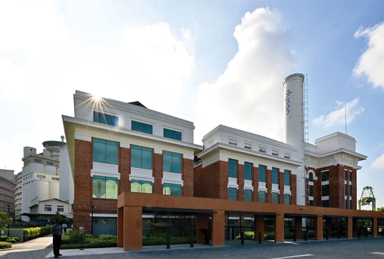 A restored red-brick heritage building with large glass windows and white trim, now serving as Dyson's headquarters at St James Power Station. A tall white chimney with the Dyson logo rises from the structure, and the sun glints off the rooftop under a blue sky.