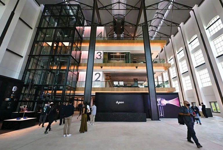 Interior of Dyson's headquarters at St James Power Station, showcasing a spacious, high-ceilinged industrial hall with glass elevators, numbered levels, and modern design elements. Visitors are seen walking through the sleek, futuristic space.