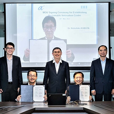 Group of business professionals pose at a conference table during an MOU signing ceremony for establishing a Sustainable Innovation Centre, with a virtual participant shown on a large screen behind them holding the signed document.