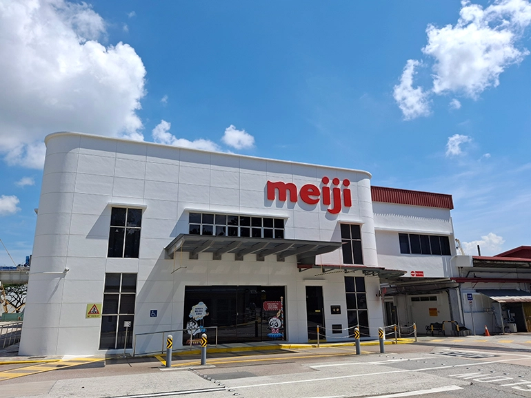 Exterior view of a Meiji facility building in Singapore, featuring the company logo on a white facade under a bright blue sky.