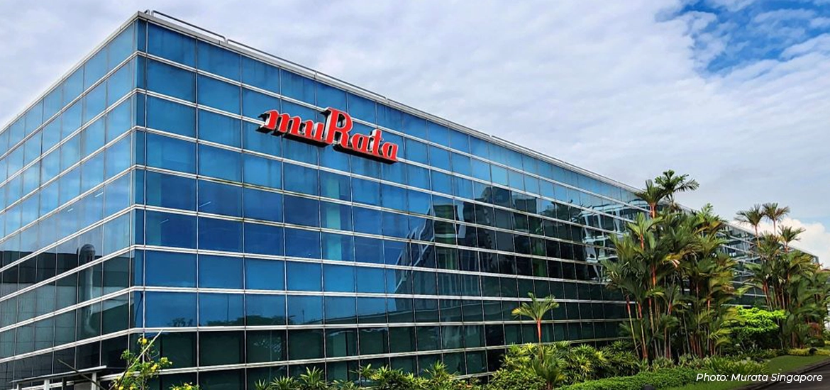 Modern glass office building with the Murata logo on the facade, surrounded by greenery under a partly cloudy sky.