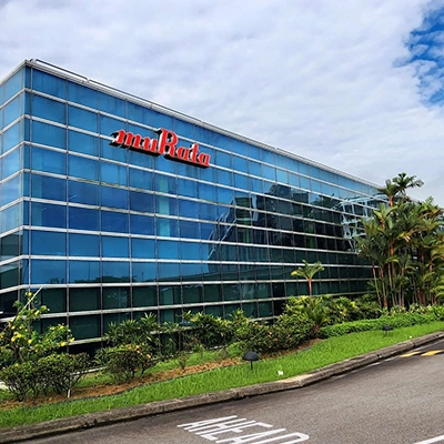 Modern glass office building with the Murata logo on the facade, surrounded by greenery under a partly cloudy sky.