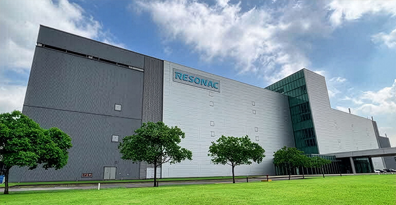 Exterior view of a Resonac manufacturing facility, featuring a modern industrial building surrounded by landscaped greenery under a bright sky.