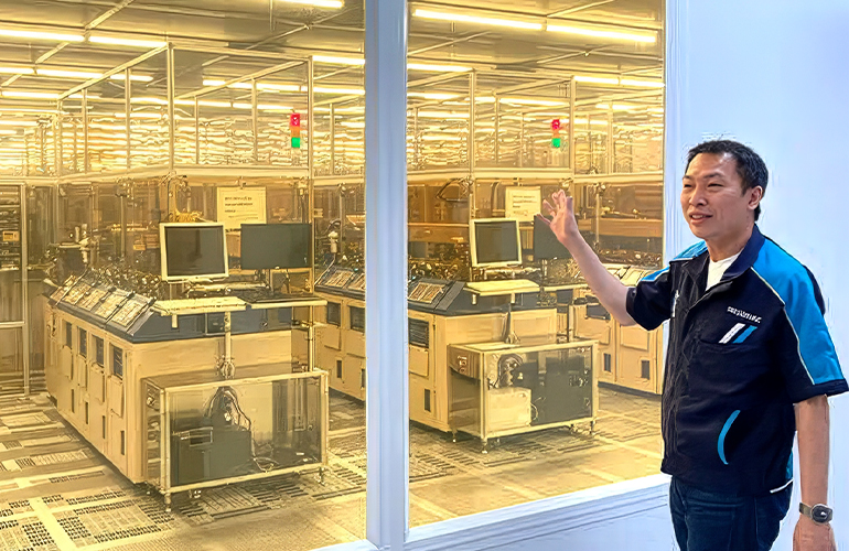 Employee standing beside a glass-enclosed semiconductor production area, presenting advanced manufacturing equipment and automated systems inside a cleanroom facility.