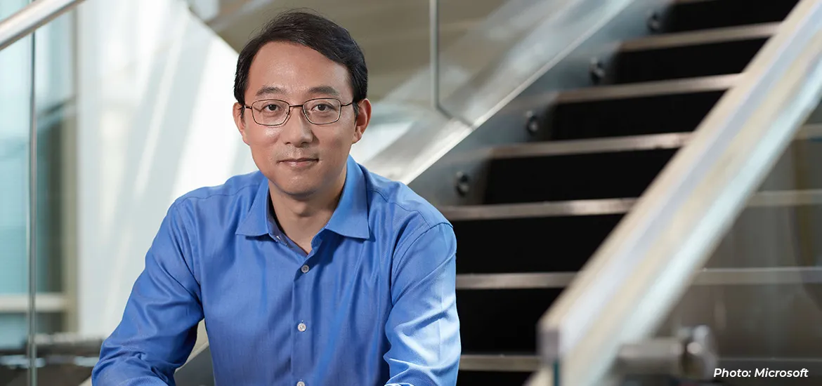 A man in a blue shirt and glasses sitting indoors near a staircase, looking at the camera in a professional setting.