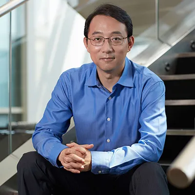 A man in a blue shirt and glasses sitting indoors near a staircase, looking at the camera in a professional setting.