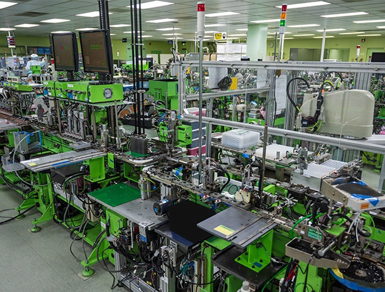 A high-tech manufacturing facility with bright lighting showcases a long, green automated production line equipped with precision machinery, control panels, and robotic arms. Multiple screens display system data, indicating a focus on automation and industrial efficiency.