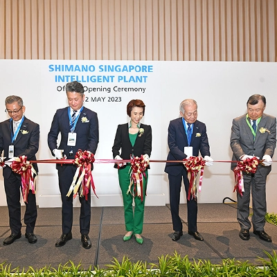 Five dignitaries stand in a row cutting a red ribbon adorned with bows at the official opening ceremony of the Shimano Singapore Intelligent Plant, held on 12 May 2023. A white banner with blue text is displayed behind them.