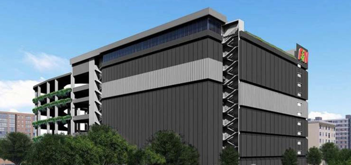 A modern, multi-story industrial building with a sleek black and grey facade, featuring vertical greenery on one side and an external staircase. The structure is labeled with the "F&N" logo on the top corner, set against a clear blue sky and surrounded by trees and nearby urban buildings.