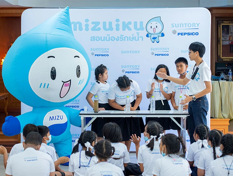 A group of children participates in an educational water conservation activity led by a presenter and accompanied by a large blue water droplet mascot named "Mizu" at a Suntory PepsiCo event. The setup includes a demonstration table and a backdrop with branding in Thai and English.