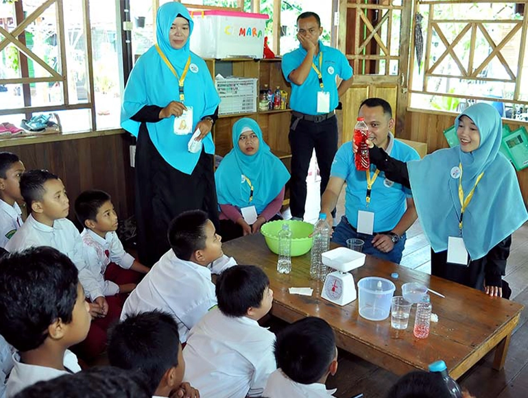 A group of children in school uniforms attentively watches a science demonstration conducted by educators wearing matching turquoise hijabs and blue shirts. The activity, held indoors in a wooden classroom, involves clear plastic bottles, measuring tools, and colored liquids.