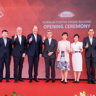 Group of executives and dignitaries posing on stage with thumbs up during the opening ceremony of Takeda’s Platinum Positive Energy Building, standing in front of a red backdrop with the Takeda logo.