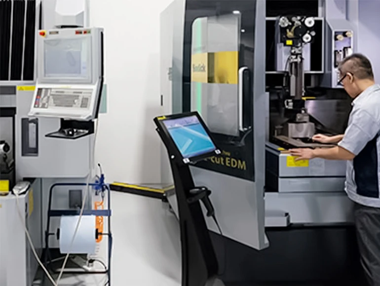 Technician operating a precision CNC or EDM machine in a clean, high-tech manufacturing facility with multiple control screens.