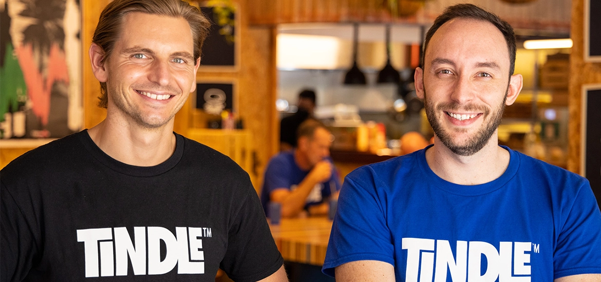 Two smiling men wearing “TiNDLE” branded t-shirts (one in black, one in blue) pose in a warmly lit restaurant setting with people dining in the background.