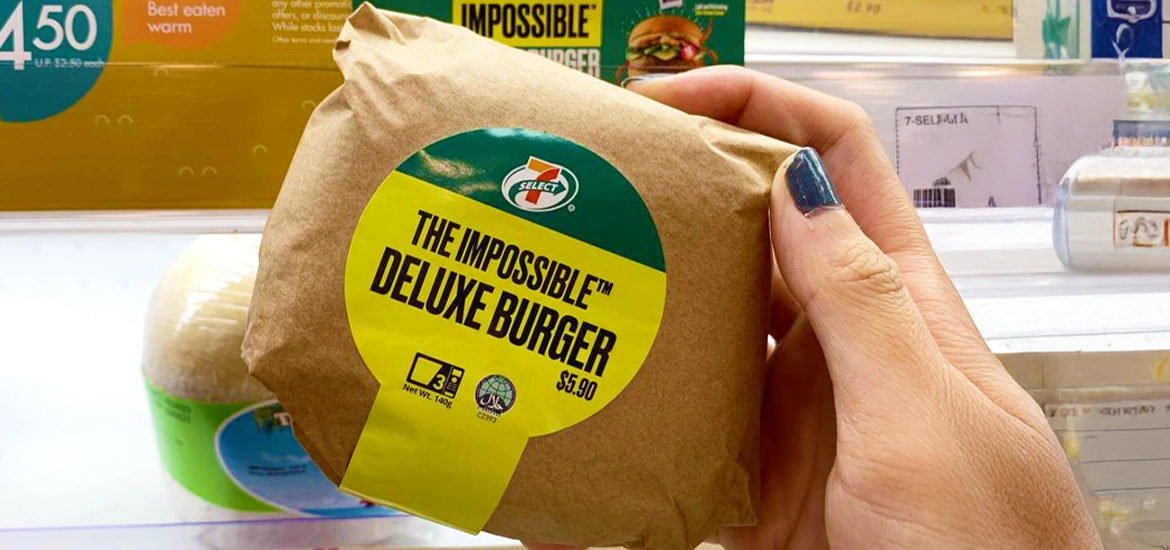 Hand holding a wrapped Impossible™ Deluxe Burger from 7-Eleven, priced at $5.90.