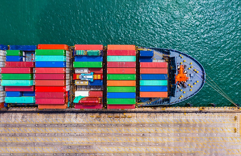 Aerial view of a cargo ship docked at a port, loaded with colorful shipping containers, beside calm blue-green water.