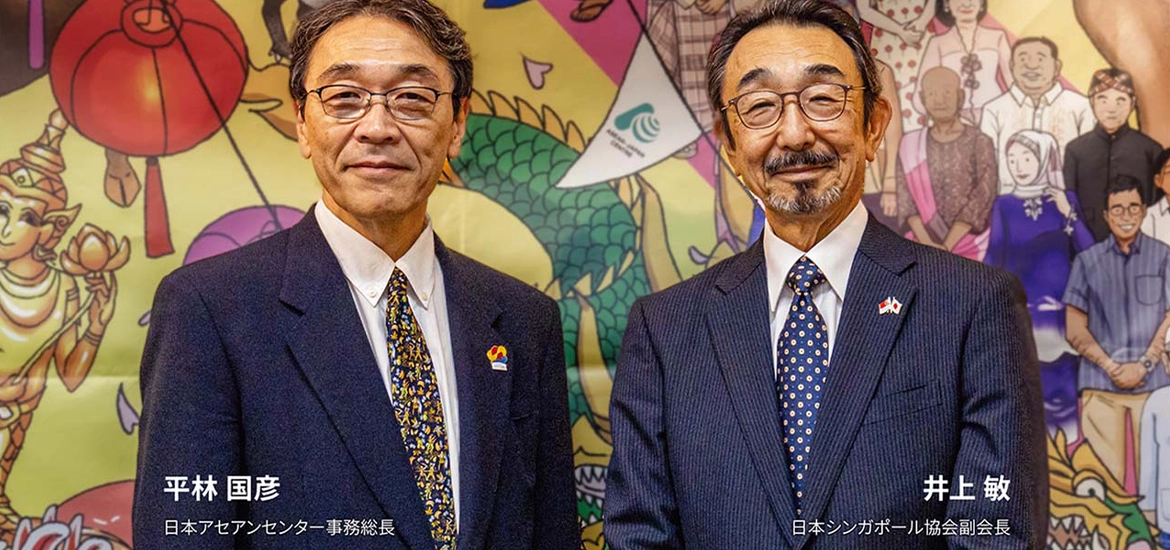 Two older men in suits smiling in front of a colorful backdrop featuring traditional Japanese-style art with dragons and vibrant elements.