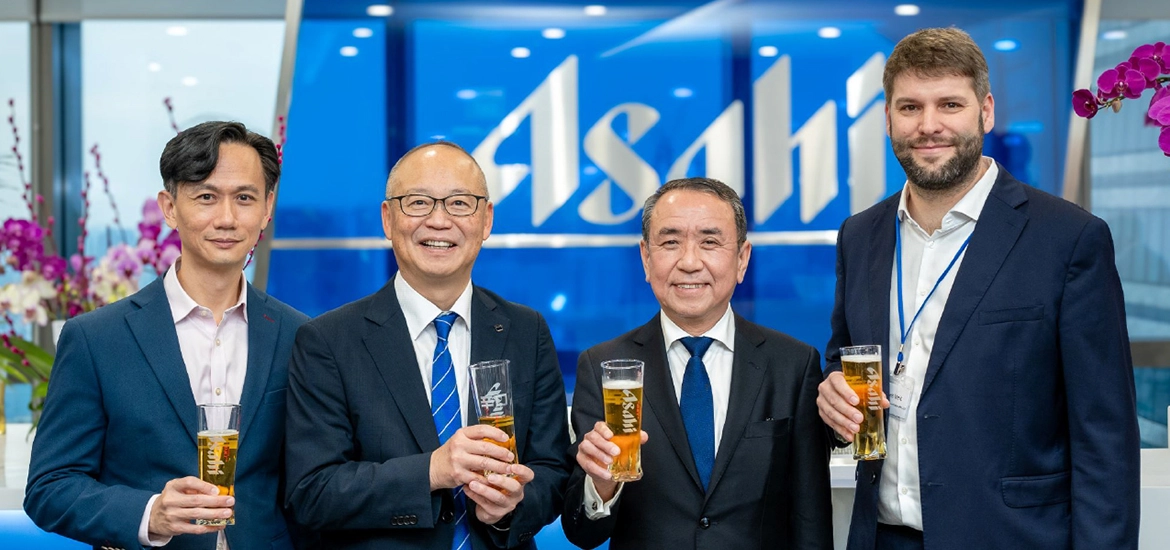 Four men in business attire smiling and holding glasses of beer in front of an Asahi logo backdrop, suggesting a corporate celebration or event.